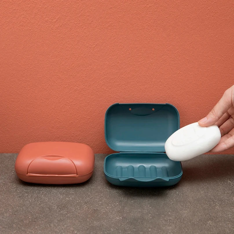 A hand opens a teal and white portable travel soap box against a terracotta-colored wall.