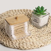 Clear glass storage box with a wooden lid holding numerous cotton swabs and pads on a woven mat.