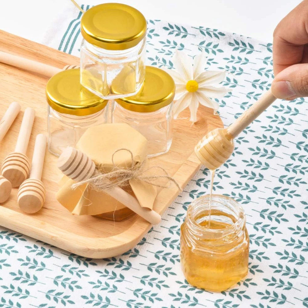 A set of mini glass honey jars with gold lids and wooden dippers displayed on a wooden board.
