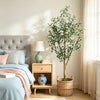 An artificial indoor olive tree in a woven basket sits on a wooden nightstand next to a bed.