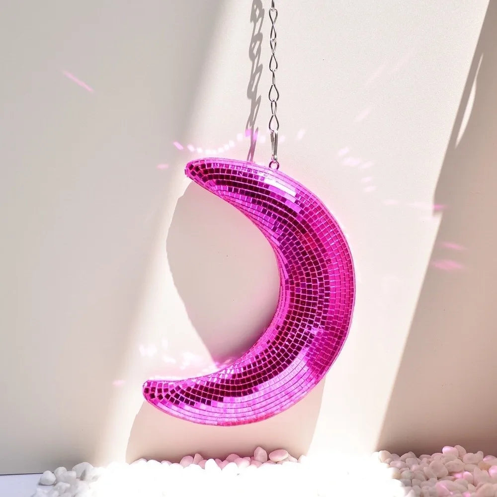 A pink iridescent mosaic moon pendant hanging against a white wall with sunlit rays.