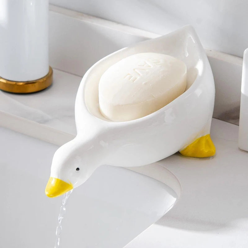 A white duck-shaped soap dish with a yellow beak is shown dripping water on a white surface.