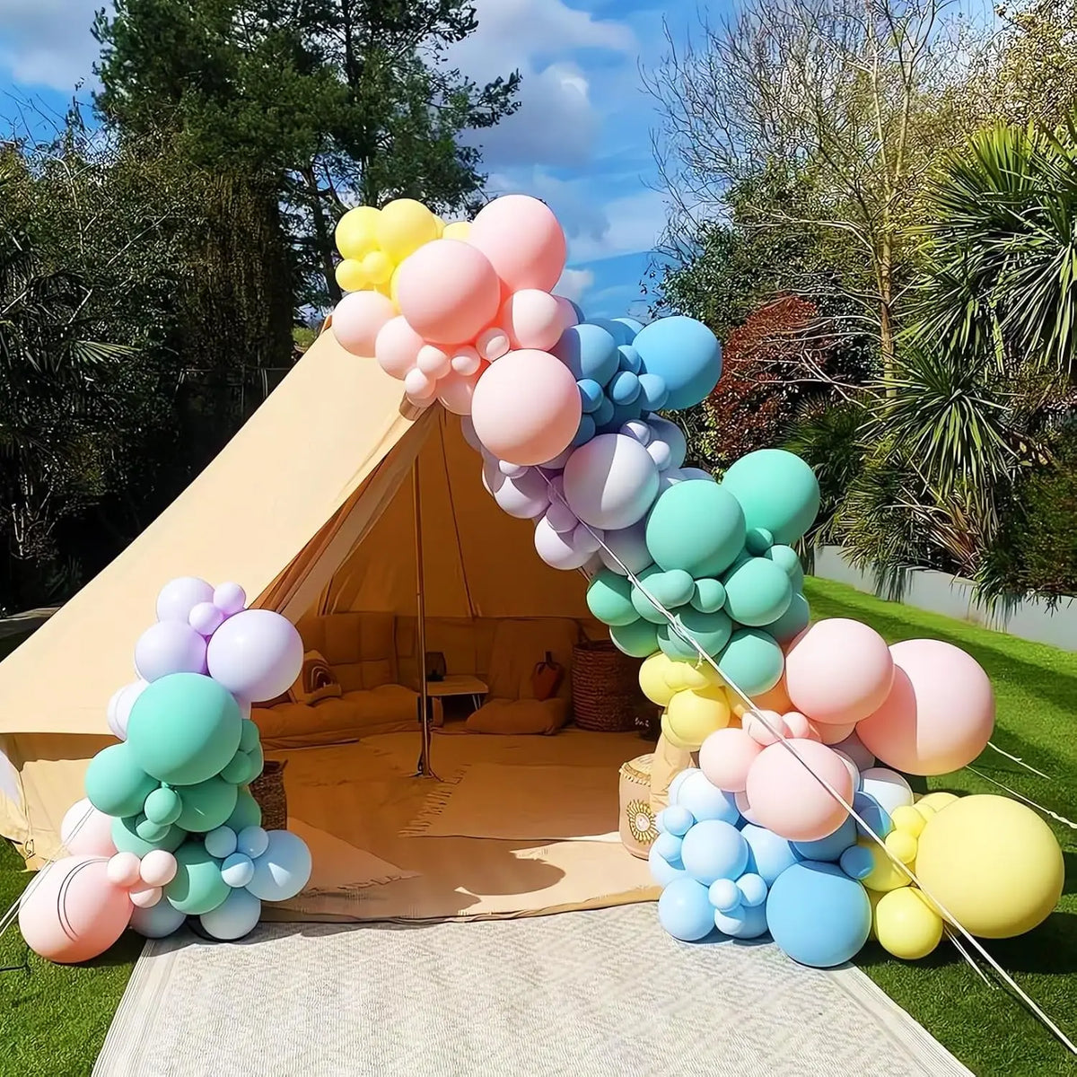 A colorful balloon arch display featuring pink, blue, and yellow balloons set up outdoors on a grassy area.