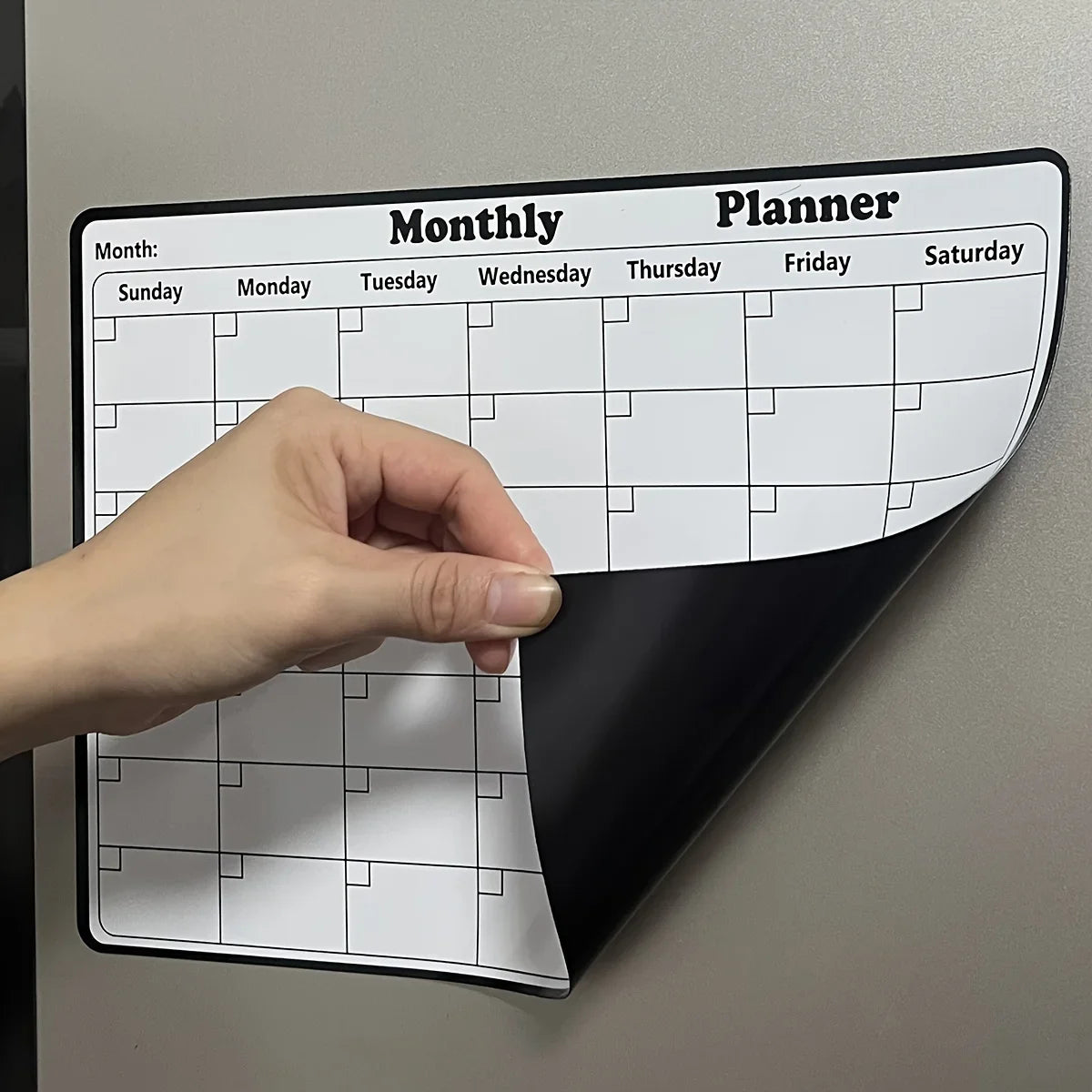 Eine Hand zeigt auf einen monatlichen Planerkalender auf dem Kühlschrank, der Wochentage und Rasterlinien zeigt.