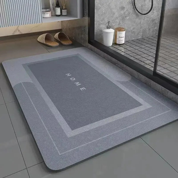 A rectangular gray anti-slip floor mat with the word HOME embossed on it placed on a tiled floor near a window.