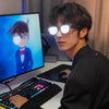 A young man wearing LED light up anime glasses is sitting at a desk with a computer monitor and keyboard.