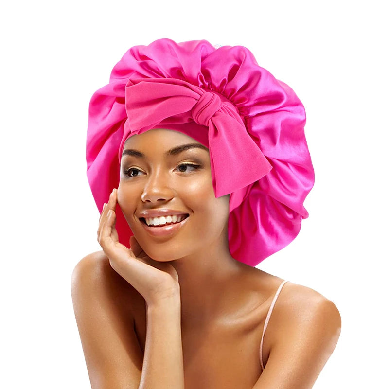 A woman wearing a bright pink satin sleeping cap tied in a bow, smiling and touching her face.