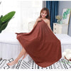 A woman models a large, rust-colored microfiber bath towel draped over her, sitting on a white bed in a bright room.