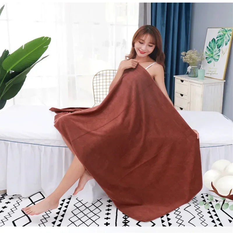 A woman models a large, rust-colored microfiber bath towel draped over her, sitting on a white bed in a bright room.