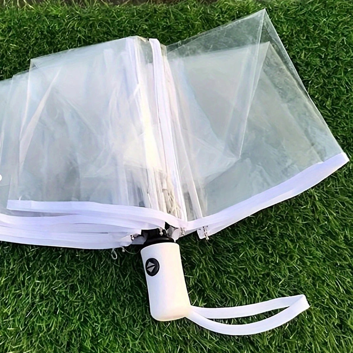 Automatic transparent folding umbrella with a white handle resting on green grass.