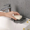 A hand is using a chrome faucet to drain a white bar of soap into a silicone draining tray on a white sink.