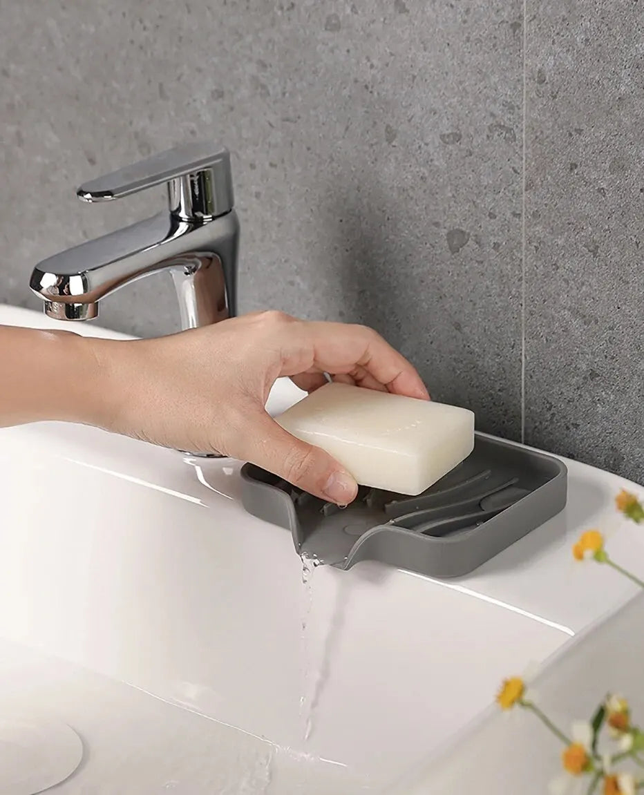A hand is using a chrome faucet to drain a white bar of soap into a silicone draining tray on a white sink.