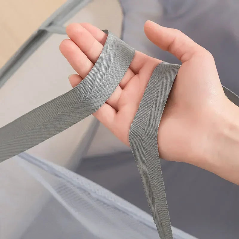 A hand is holding a gray fabric strap, which is part of a foldable mesh laundry basket.