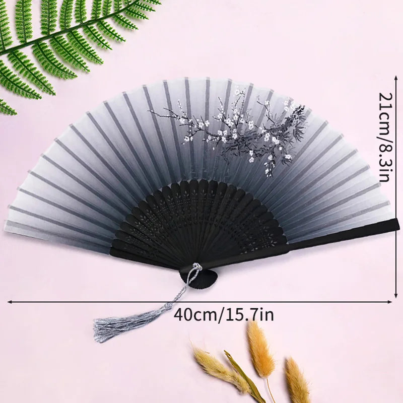 A black and gray lace fabric folding hand fan with delicate floral accents shown against a pink background.