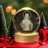 A glass crystal ball with a depiction of Jesus, set on a wooden base, surrounded by green pine trees and red ribbon.