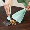 A hand is using a small green broom and dustpan set to clean a dark wooden surface.