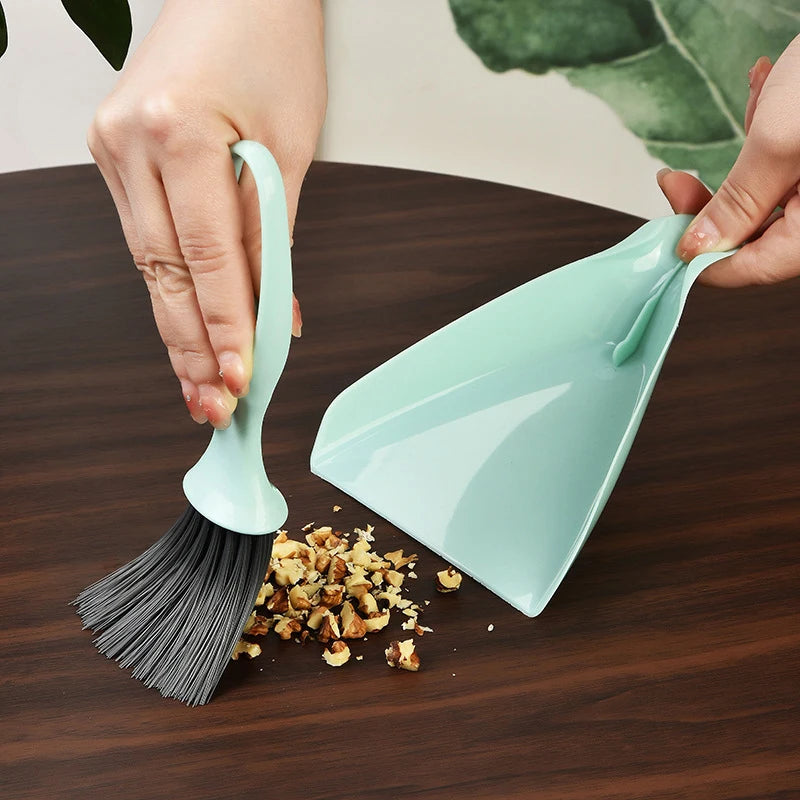 A hand is using a small green broom and dustpan set to clean a dark wooden surface.