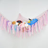 A colorful bohemian stuffed animal hammock featuring pink, blue, and orange plush toys hanging against a white wall.