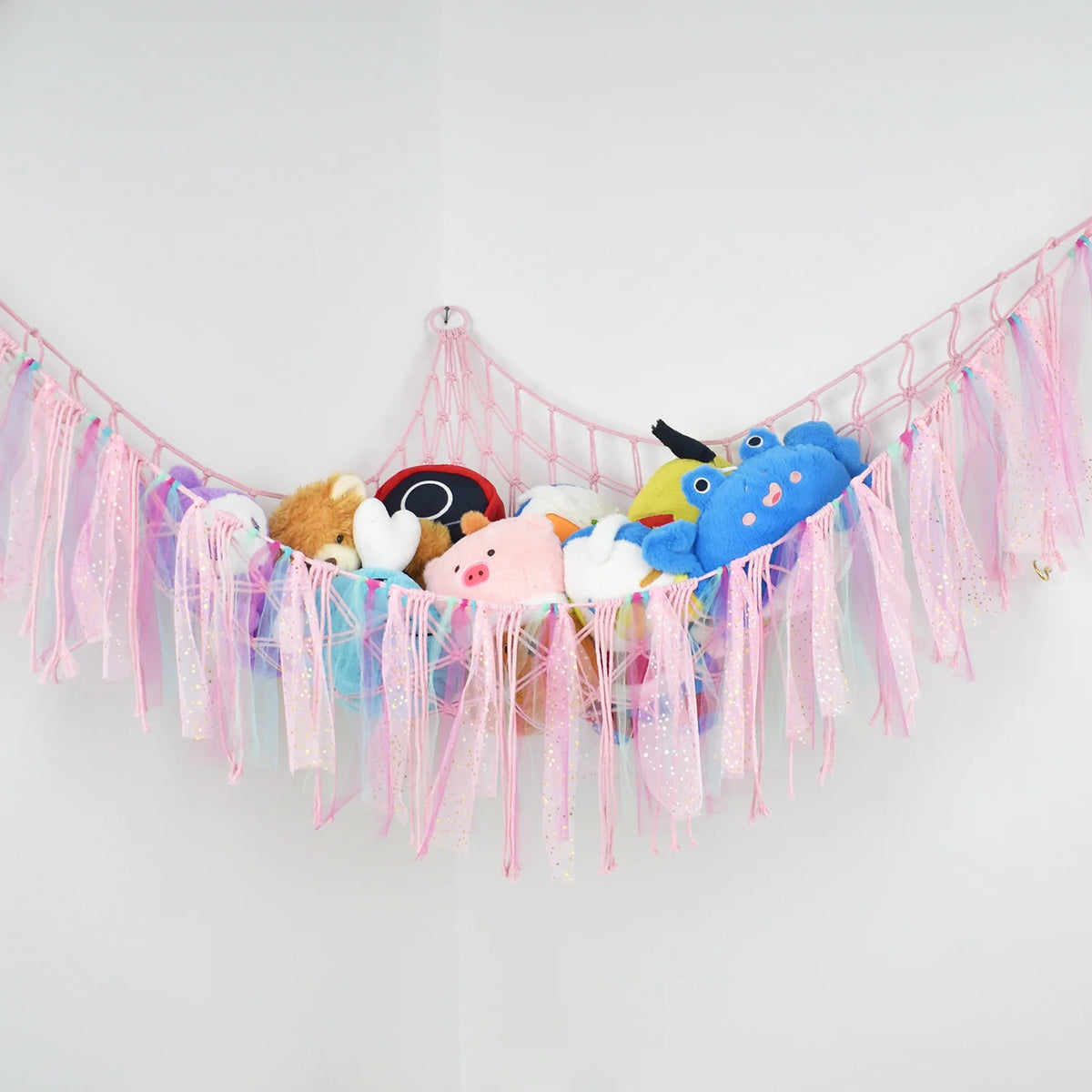 A colorful bohemian stuffed animal hammock featuring pink, blue, and orange plush toys hanging against a white wall.