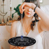 A woman holds a black divination circle with a silver pendant and a crystal necklace against a white background.