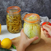 Hands are squeezing a large glass mason jar filled with sliced cucumbers and a lemon on a wooden cutting board.