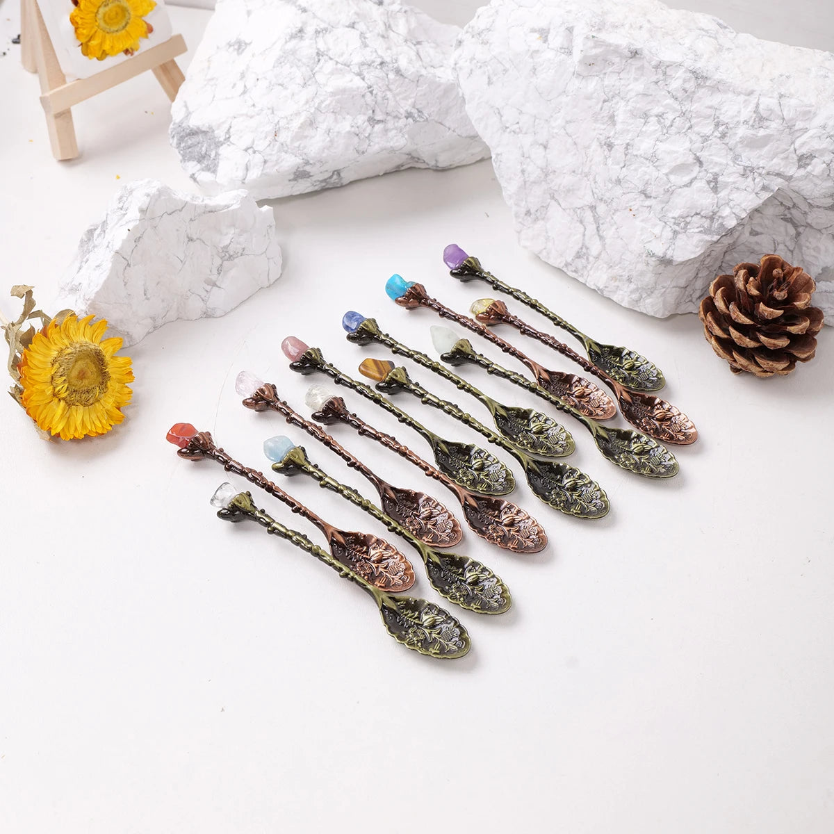 A collection of natural crystal spoons with various colors and textures arranged on a white surface next to rocks and a pinec