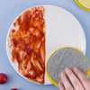 A hand is holding a yellow and gray reusable double-sided dishwashing sponge set on a white plate with a red sauce.