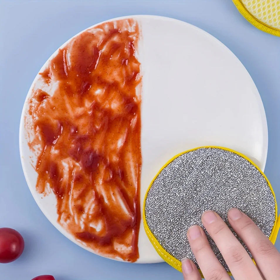 A hand is holding a yellow and gray reusable double-sided dishwashing sponge set on a white plate with a red sauce.