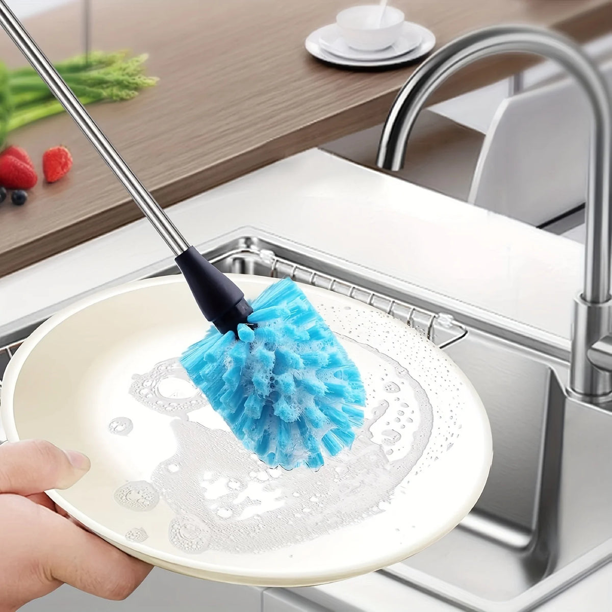 A hand is using a blue sponge brush on a white plate in a kitchen sink to clean dishes.