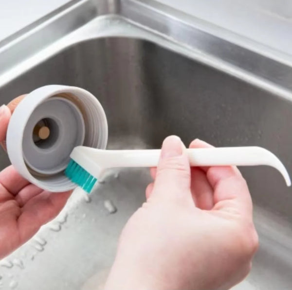 A hand is using a white silicone cleaning brush to clean a bottle inside a stainless steel sink.
