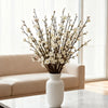 A vase filled with artificial plum blossoms on a glass table near a window.