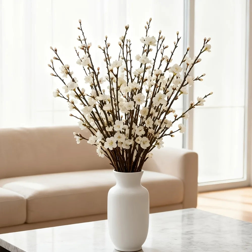 A vase filled with artificial plum blossoms on a glass table near a window.