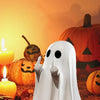 A white ghost statue decoration stands in front of carved pumpkins and a glowing candle on a dark surface.