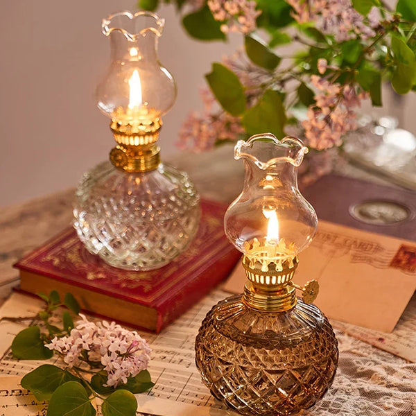 A vintage rustic oil lamp in glass and brass sits on a wooden surface next to a book and floral elements.