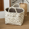 A cream-colored quilted storage basket with a handle and floral pattern sits on a wooden floor next to a potted plant.