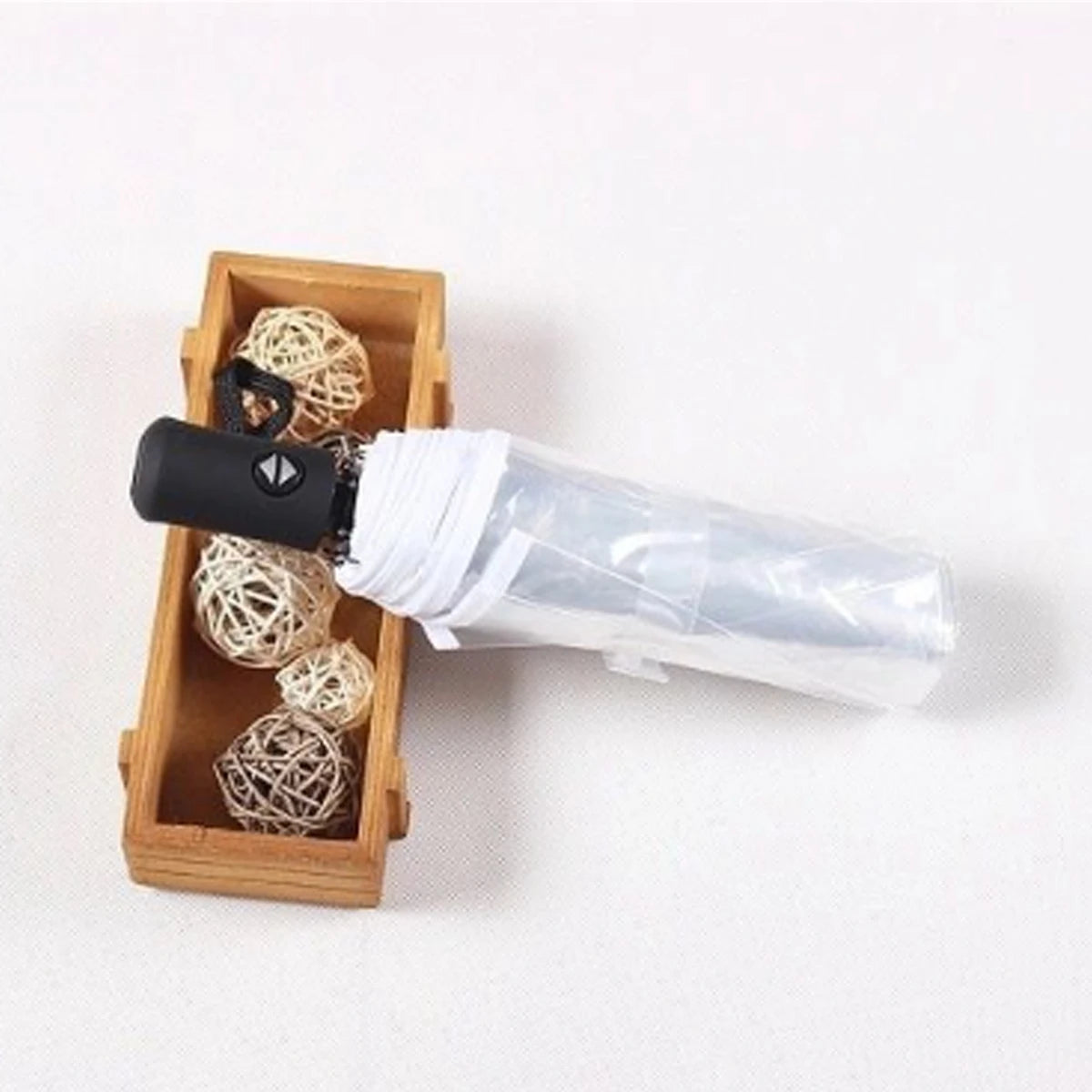 A black automatic transparent folding umbrella is shown next to a wooden box containing several small, spherical, light-color