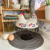 A white ceramic hen-shaped fruit basket sitting on a dark woven mat next to a wooden shelf.