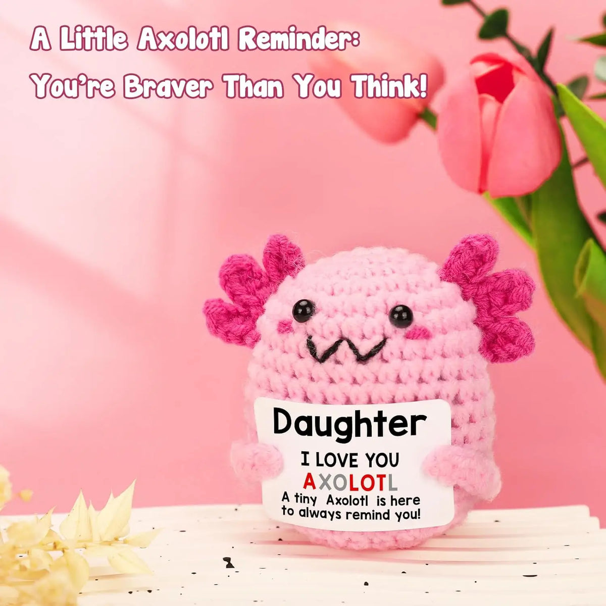 A pink handmade crochet axolotl figurine with pink frills sits on a white surface.