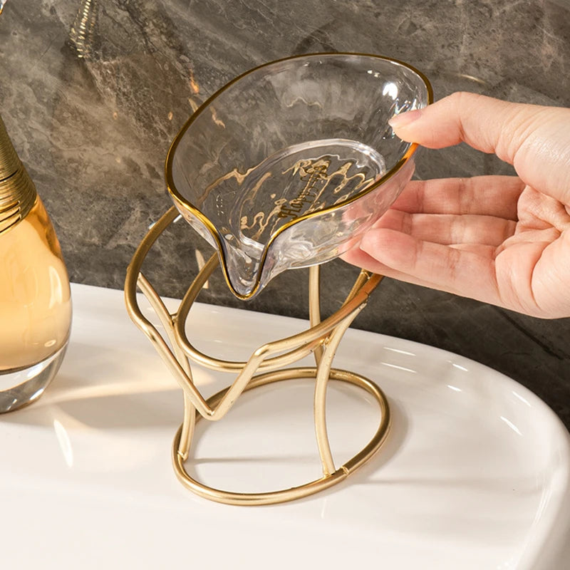 A hand holds a gold-toned decorative leaf soap dish made of clear glass, resting on a white tray.