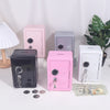 A collection of solid color metal piggy banks in pink, gray, and black, with a white lamp and US currency nearby.