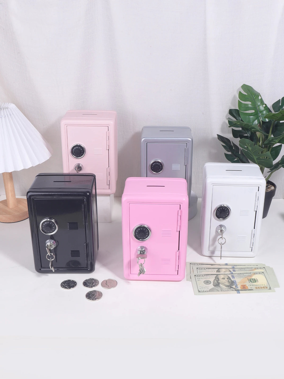A collection of solid color metal piggy banks in pink, gray, and black, with a white lamp and US currency nearby.