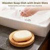 A wooden soap dish with drain slots is shown next to a bar of natural soap being washed in a white sink.
