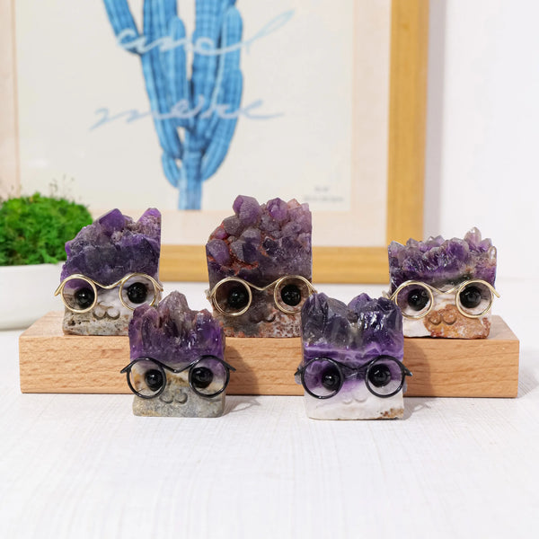 Five natural amethyst crystal wear glasses displayed on a wooden surface with a blurred blue cactus in the background.