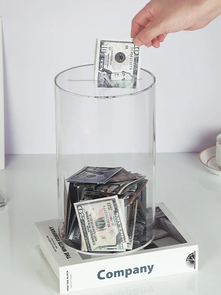 Una mano coloca una pila de billetes de dólar en una alcancía de acrílico transparente junto a otra moneda.