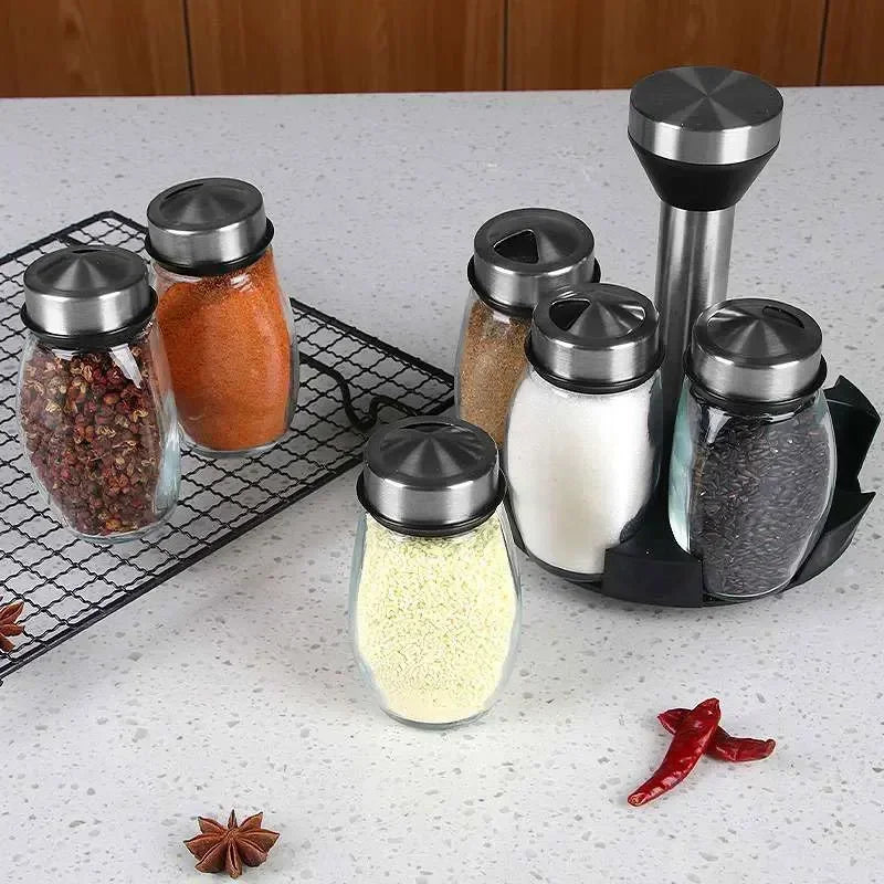 A set of rotating glass spice jars with stainless steel lids and a stand, displaying various ground spices on a light surface