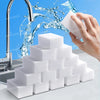 A hand is squeezing a white melamine foam cleaning sponge under running water, with a stack of similar sponges in the foregro