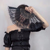A woman holds a black lace fabric folding hand fan with intricate detailing against a light gray background.
