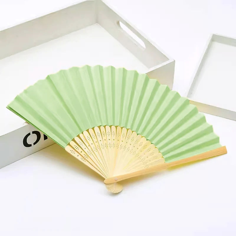 Un éventail pliable vert clair en bambou et papier, ouvert sur fond blanc.