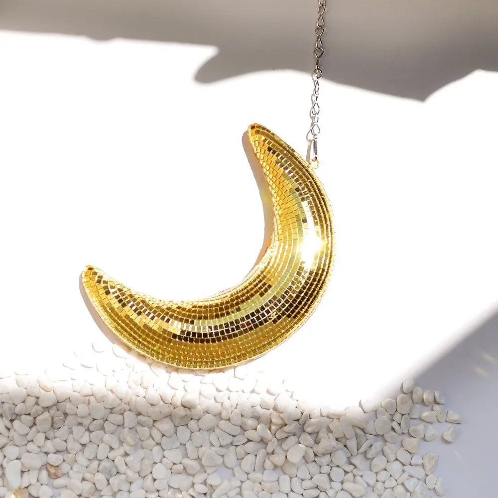 A gold glitter moon pendant hanging from a silver chain, resting on a bed of small white pebbles.
