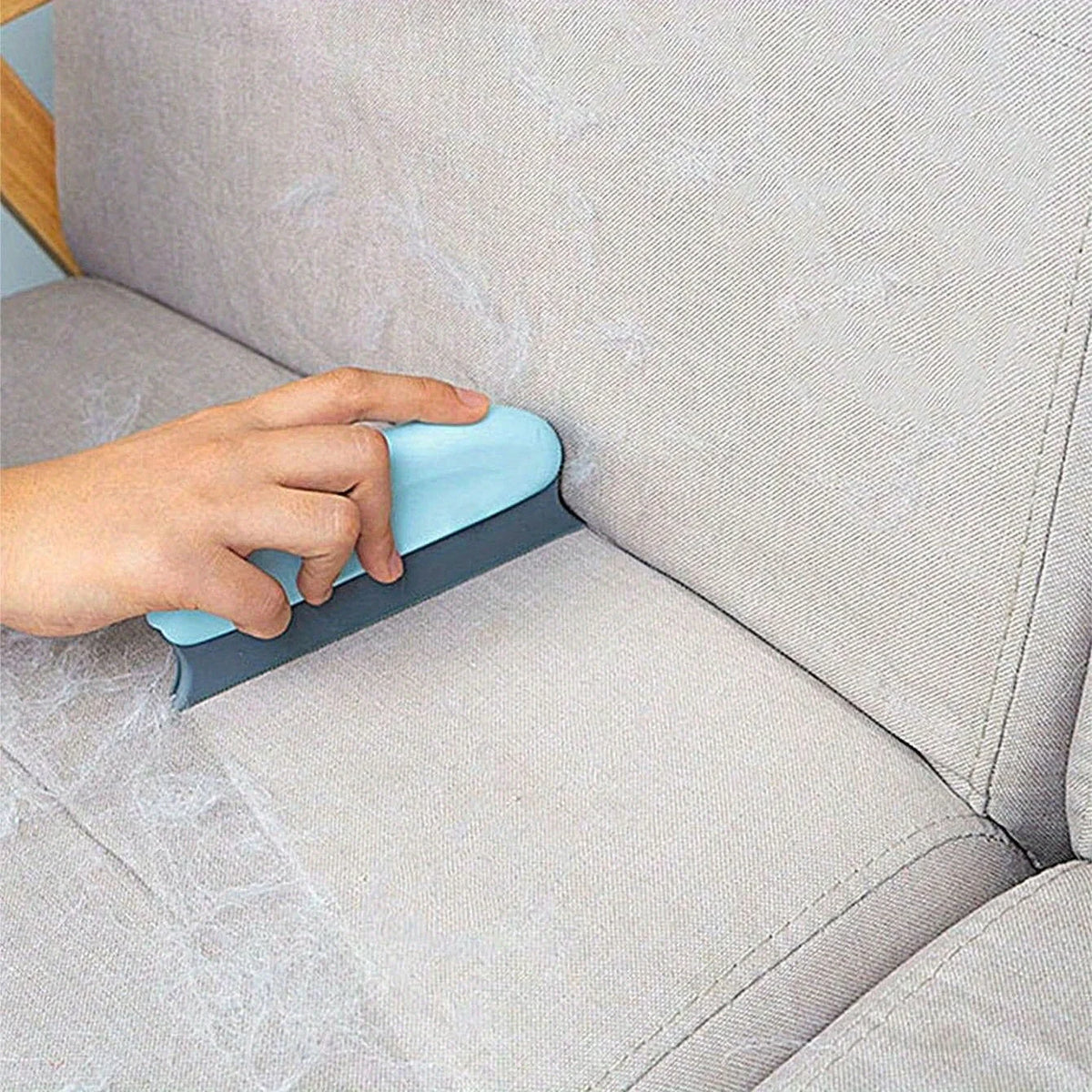 A hand is using a blue pet hair remover brush on a light gray sofa fabric.